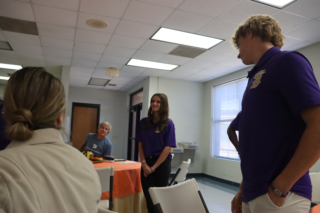A Bearcat Ambassador student stands to introduce herself during the meeting while another student stands nearby. Adults seated at tables listen attentively.
