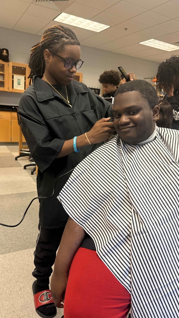 A student barber with locs and glasses gives a haircut to a student sitting in a striped cape. The student smiles while the barber focuses on using clippers. Other students work in the background.
