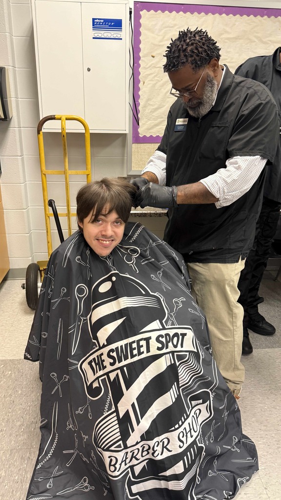 A barber with short twists and a gray beard trims a smiling student’s hair in a classroom. The student sits under a black barber cape labeled “The Sweet Spot Barber Shop.” The atmosphere is warm and friendly.