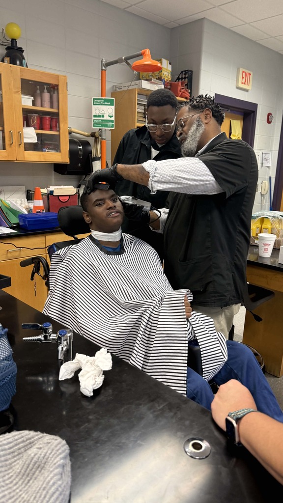 A student in a wheelchair receives a haircut from an adult barber while another student barber observes closely. The student wears a striped cape and looks upward as the haircut continues.
