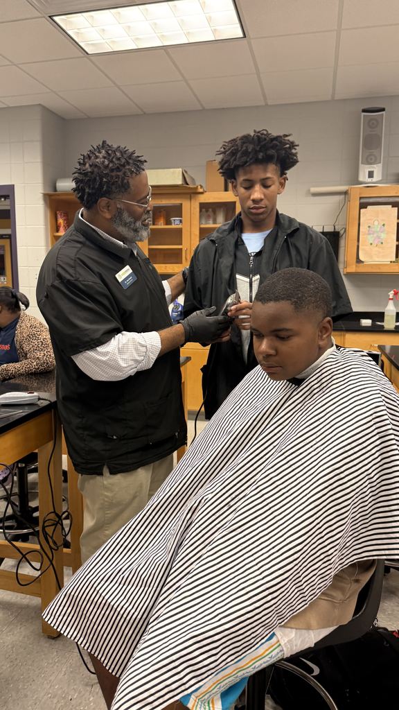 An adult barber teaches a student barber how to use clippers while giving a haircut to a boy wearing a striped cape. The student in the chair looks calm and focused during the demonstration.
