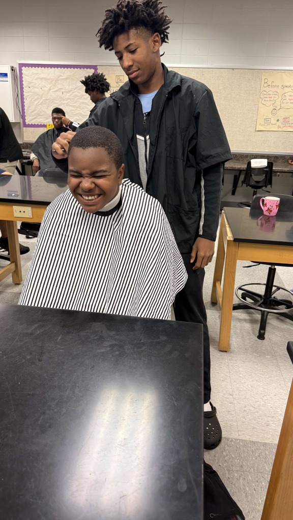 A student barber carefully trims another student’s hair. The student receiving the haircut is smiling widely with his eyes closed, seated at a classroom table and wearing a striped barber cape.