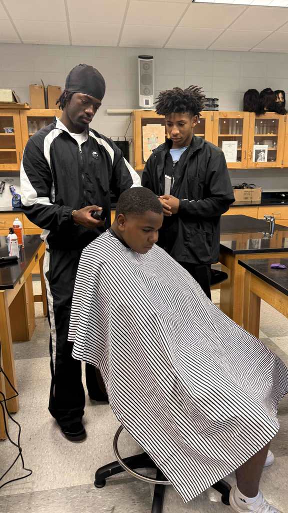 A student barber and another student observer stand beside a classmate getting a haircut in the classroom. The seated student wears a striped cape while the barber trims the sides of his hair with clippers.
