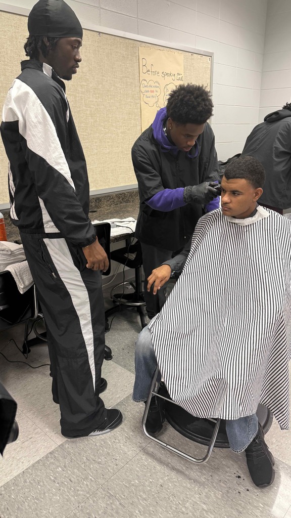 Two student barbers stand beside a classmate seated in a chair while one trims his hair. The student receiving the haircut wears a striped cape and looks straight ahead as the others assist and observe.