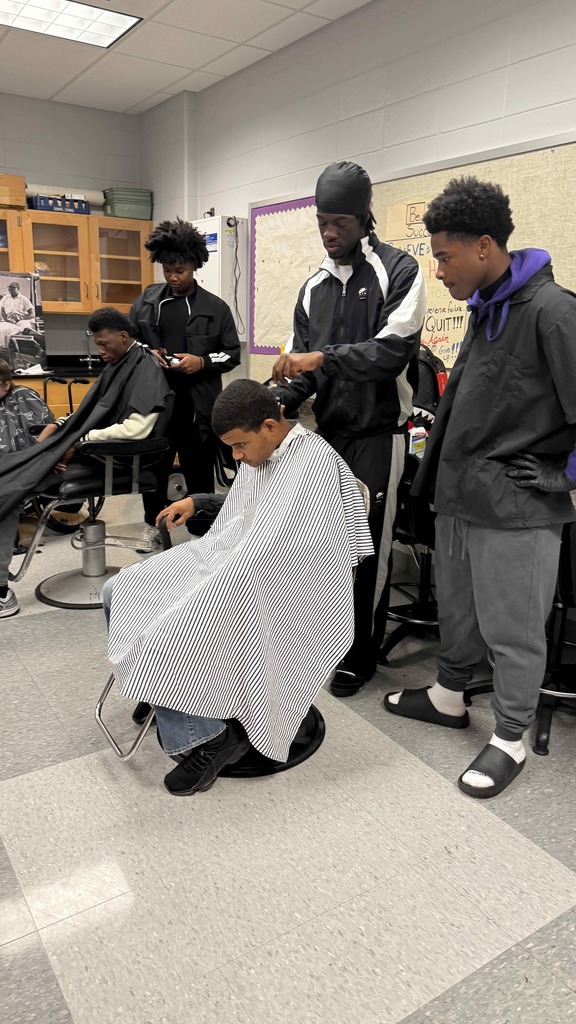 Several student barbers stand around a classmate getting a haircut. One student is actively trimming while the others observe closely. The student in the chair wears a striped cape and looks downward.