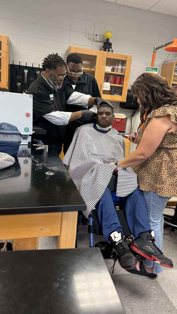 An adult barber and a student barber work together to give a haircut to a student seated in a wheelchair. A teacher stands nearby assisting. The student wears a striped barber cape and looks forward.