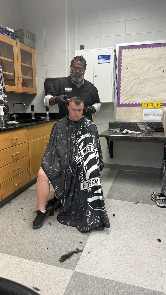 An adult barber gives a haircut to a student with Down syndrome. The student sits still in a black “Sweet Spot Barber Shop” cape while trimmed hair falls onto the floor around the chair.