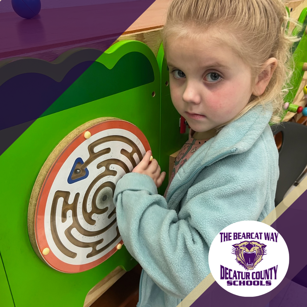 A young girl in a light blue jacket plays with a circular maze activity board at school, looking toward the camera. A Decatur County Schools “The Bearcat Way” logo appears in the corner.