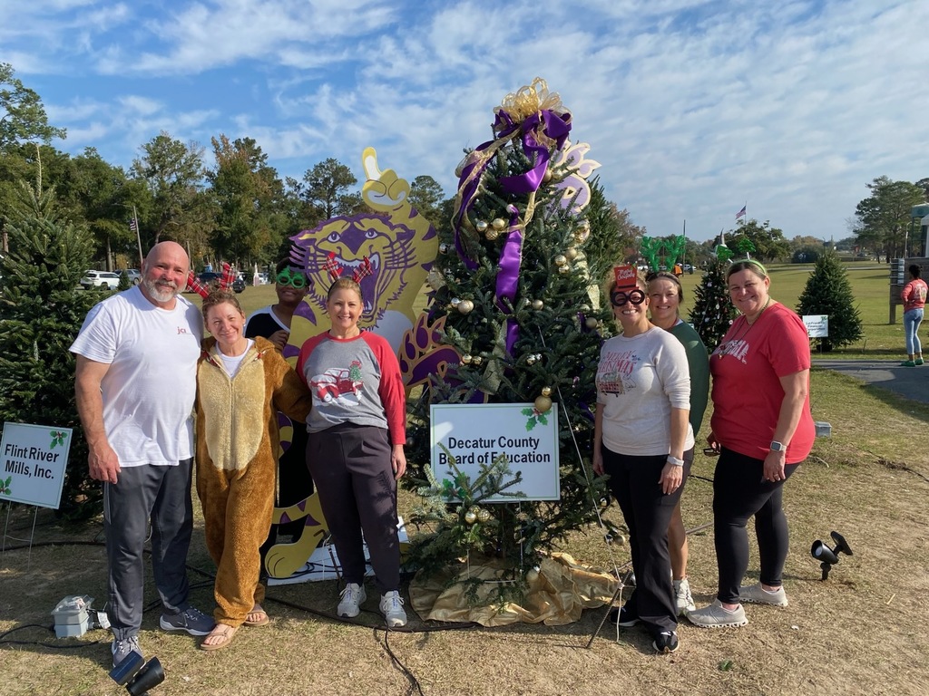 A group of six smiling adults stands outdoors beside a decorated Christmas tree labeled “Decatur County Board of Education,” with festive props and additional trees in the background.