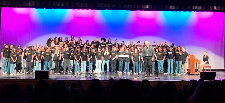 A picture of all three grade levels of Chorus. They are standing on stage with a blue and purple light effect behind them.