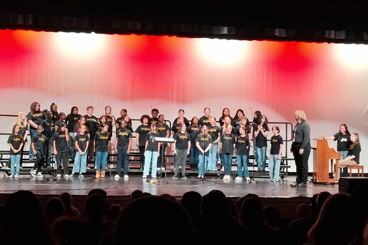 6th Grade Chorus students standing on stage with a red and white lighting effect behind them.