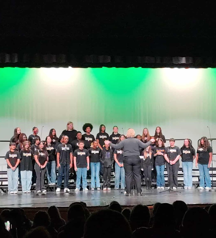 8th grade chorus students standing on stage with a green and white lighting effect behind them.