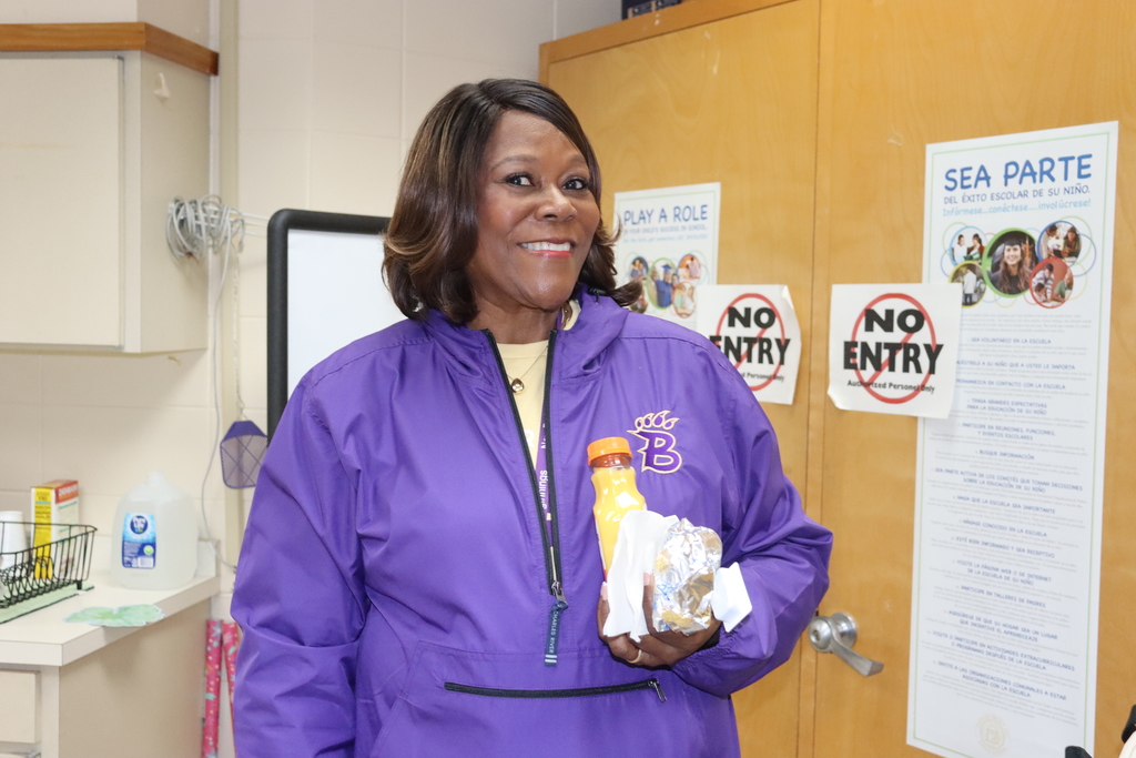 A staff member wearing a purple jacket and holding a biscuit and orange juice smiles while standing near a classroom door and informational posters.