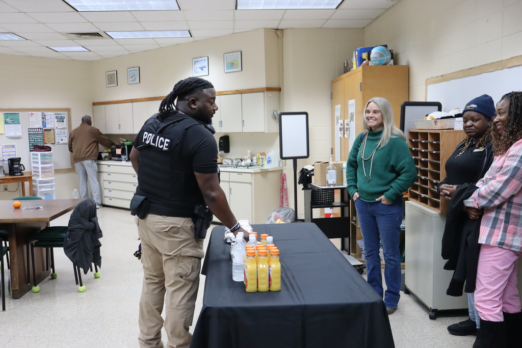 A police officer stands at a table with breakfast items, speaking to three smiling staff members in a breakroom. Another person is preparing food in the background.