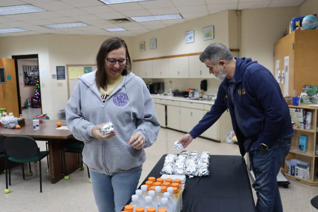 A staff member wearing an NBLC hoodie picks up a foil-wrapped biscuit while another staff member arranges more breakfast items on the table beside her.