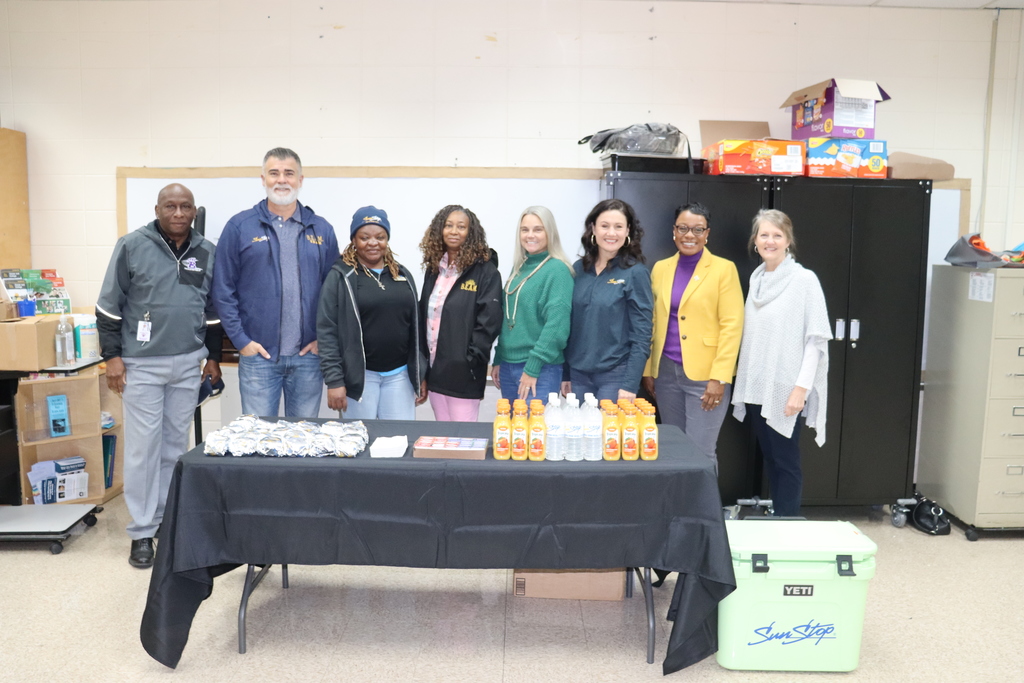 A group of nine school staff members stand behind a table filled with wrapped breakfast biscuits, juices, and water bottles in a breakroom setting.