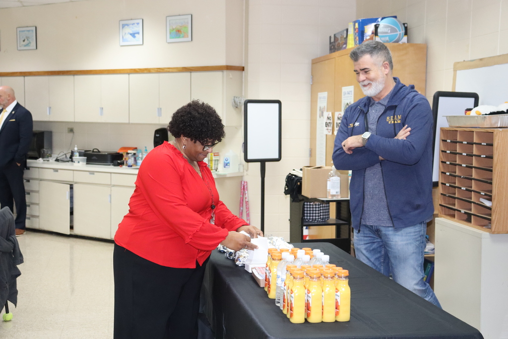 A staff member in a red top selects a foil-wrapped biscuit from a table of breakfast items while another staff member stands nearby smiling with arms crossed.