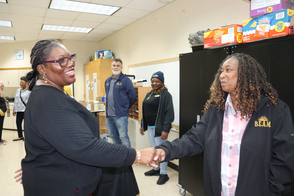 Two staff members greet each other with a friendly fist bump in a breakroom while others smile and observe in the background.