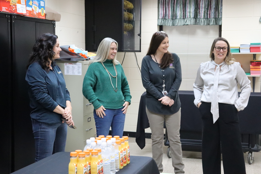 Four staff members stand together and smile during a breakfast event, with a table of juices and water bottles visible in the foreground.