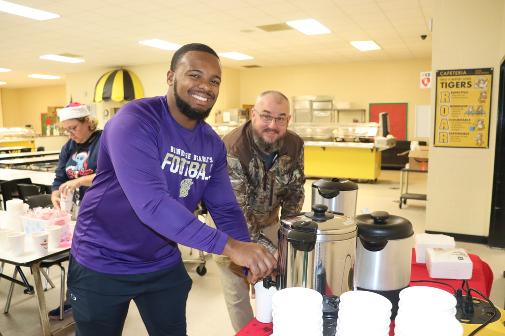 Two adults smile while serving hot cocoa from large drink dispensers in a cafeteria.