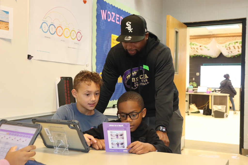 Adult mentor helps two students follow coding instructions on a tablet at a classroom table.