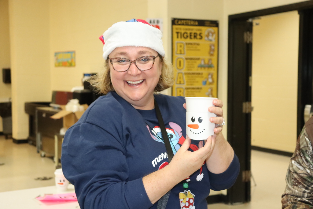 Smiling adult wearing a Santa hat holds a snowman-themed cup indoors.