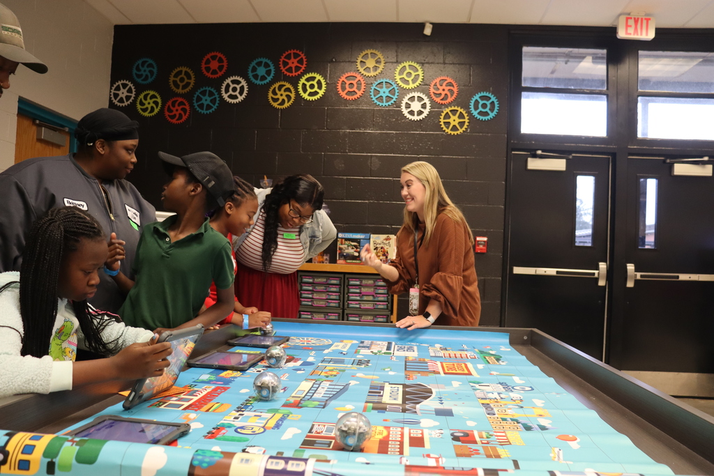 Students gather around a large robot coding mat while an adult facilitator gives instructions