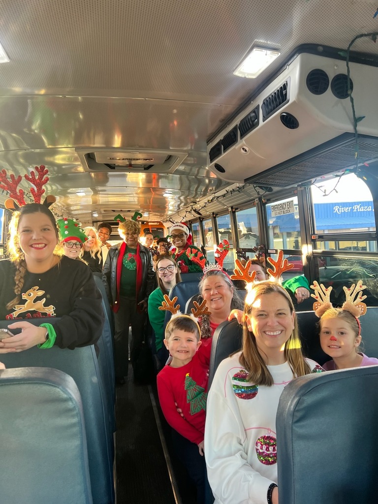 Smiling adults and children wearing festive holiday outfits and reindeer antlers seated on a school bus.