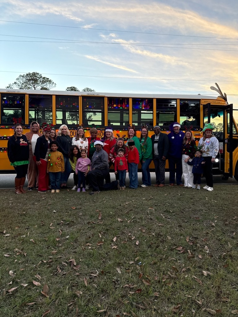 Group of adults and children posing in front of a school bus decorated with holiday lights at dusk.
