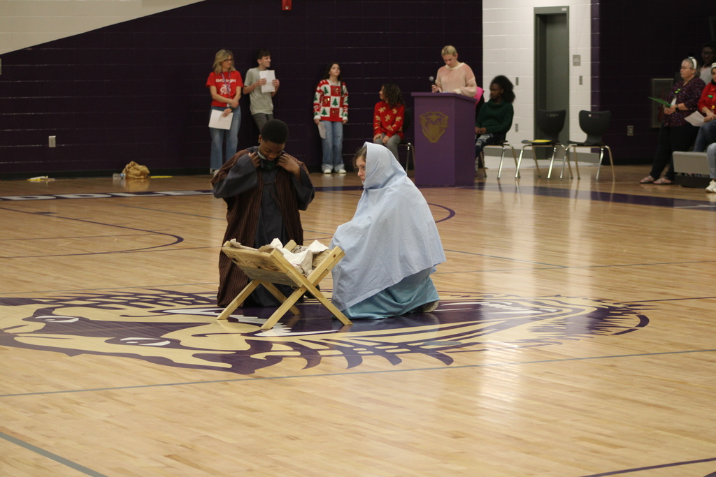 Bainbridge Middle School students participate in a nativity program in the school gym, wearing costumes, kneeling, and standing together as classmates and staff observe.
