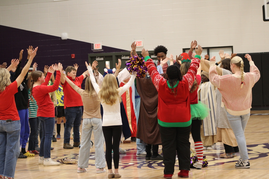 Bainbridge Middle School students participate in a nativity program in the school gym, wearing costumes, kneeling, and standing together as classmates and staff observe.