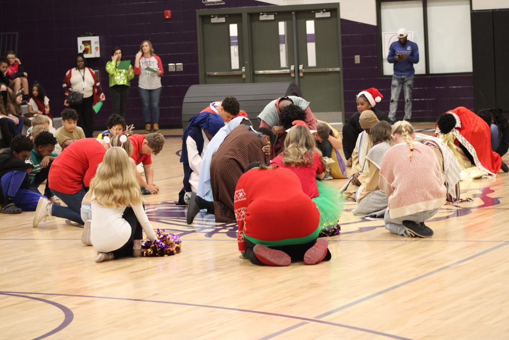 Bainbridge Middle School students participate in a nativity program in the school gym, wearing costumes, kneeling, and standing together as classmates and staff observe.