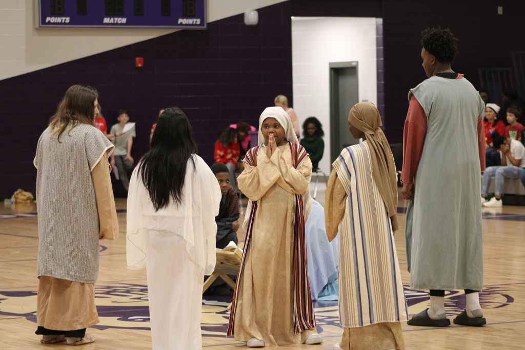 Bainbridge Middle School students participate in a nativity program in the school gym, wearing costumes, kneeling, and standing together as classmates and staff observe.