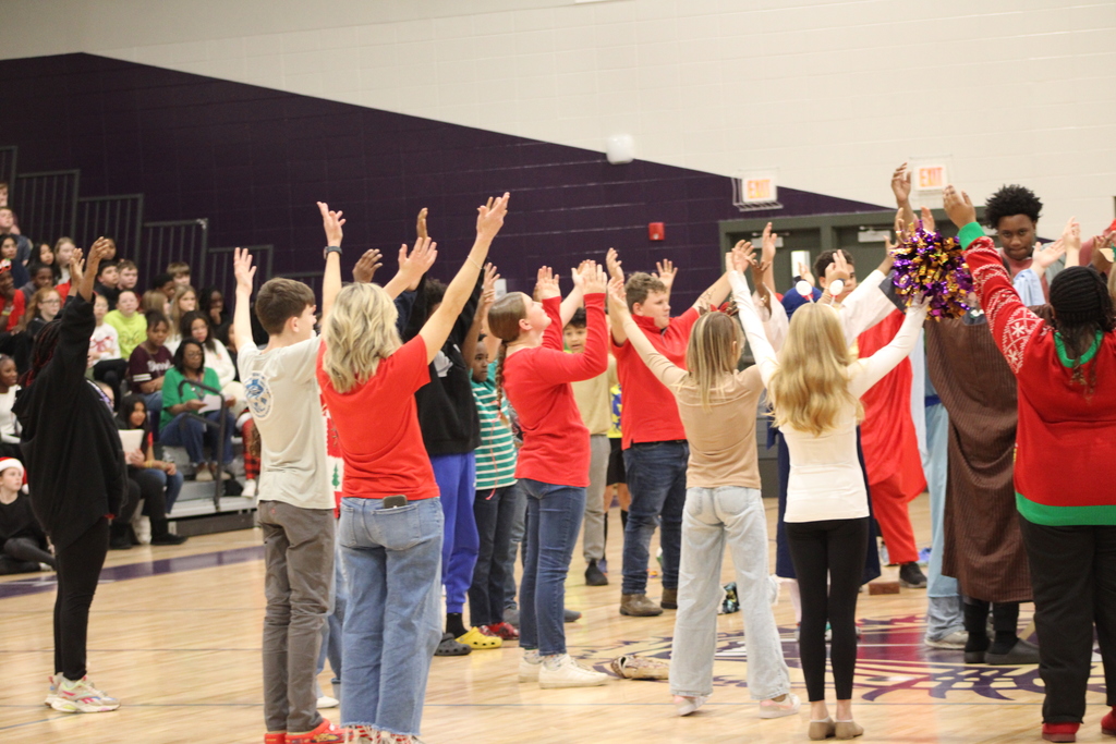 Bainbridge Middle School students participate in a nativity program in the school gym, wearing costumes, kneeling, and standing together as classmates and staff observe.