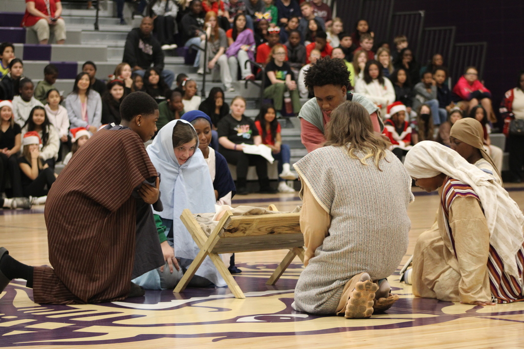 Bainbridge Middle School students participate in a nativity program in the school gym, wearing costumes, kneeling, and standing together as classmates and staff observe.