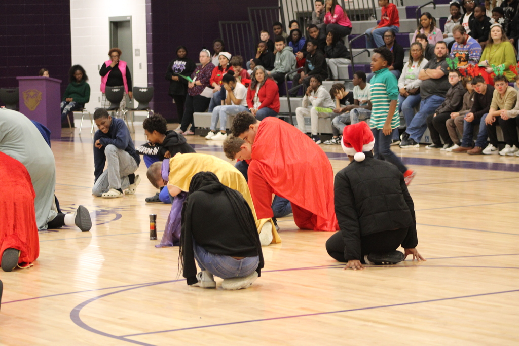 Bainbridge Middle School students participate in a nativity program in the school gym, wearing costumes, kneeling, and standing together as classmates and staff observe.