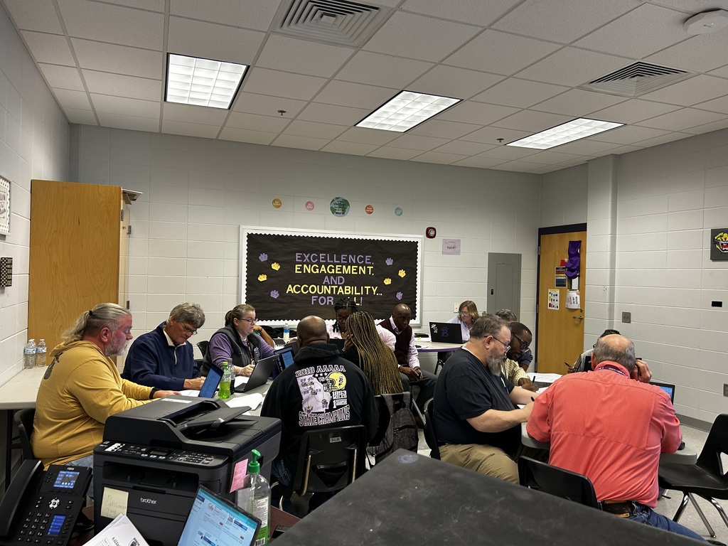 A group of educators sit around a table in a classroom, collaborating and discussing ideas during a professional meeting, with laptops open and a presentation displayed on a screen in the background.