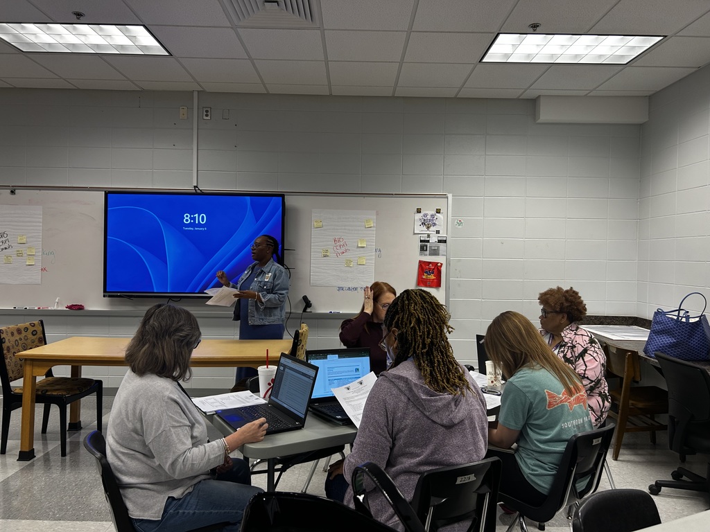 A group of educators sit around a table in a classroom, collaborating and discussing ideas during a professional meeting, with laptops open and a presentation displayed on a screen in the background.