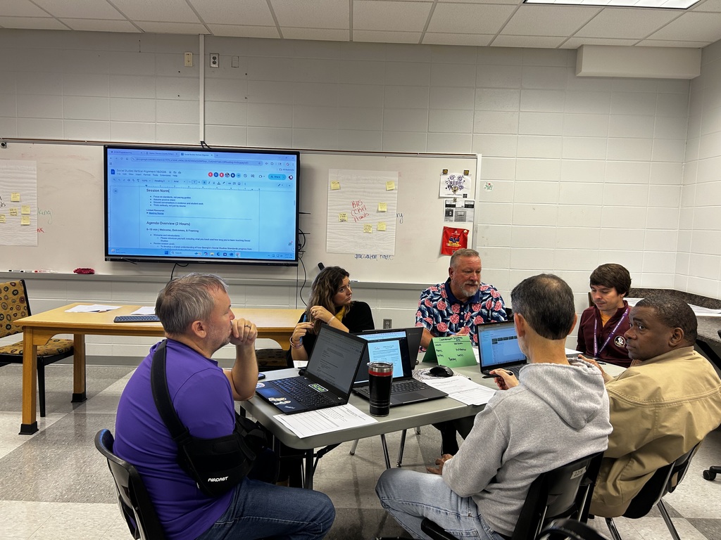 A group of educators sit around a table in a classroom, collaborating and discussing ideas during a professional meeting, with laptops open and a presentation displayed on a screen in the background.