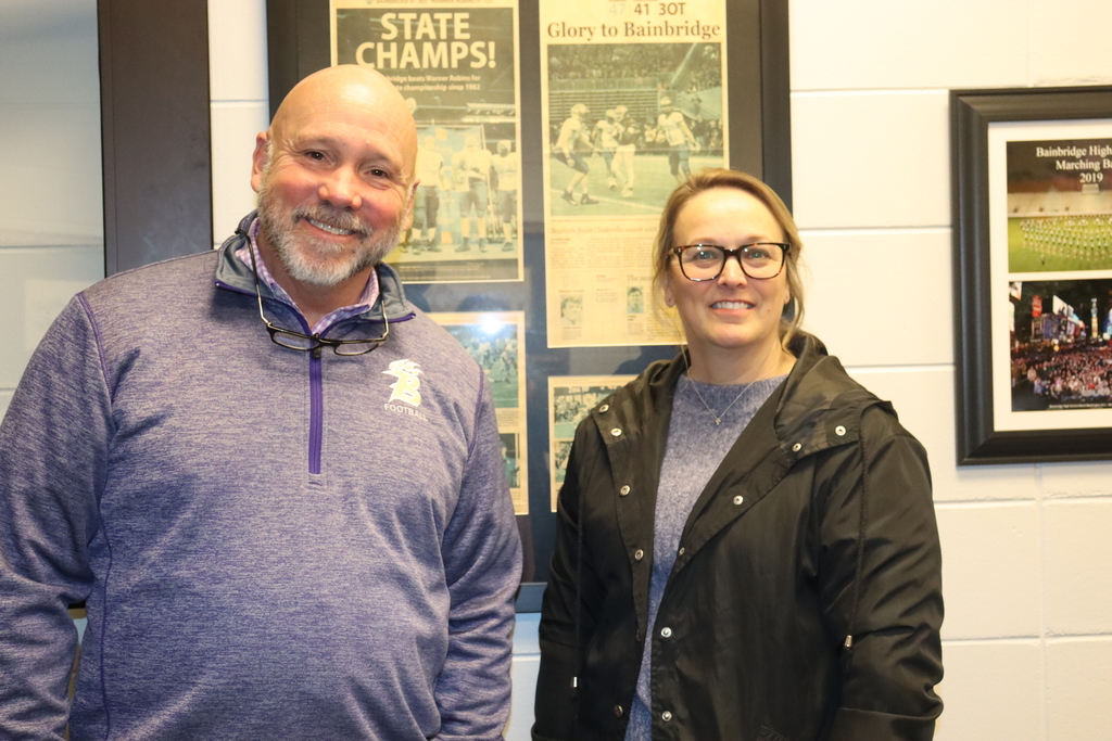 Two Bainbridge High School staff members smile while standing in front of framed school sports memorabilia.