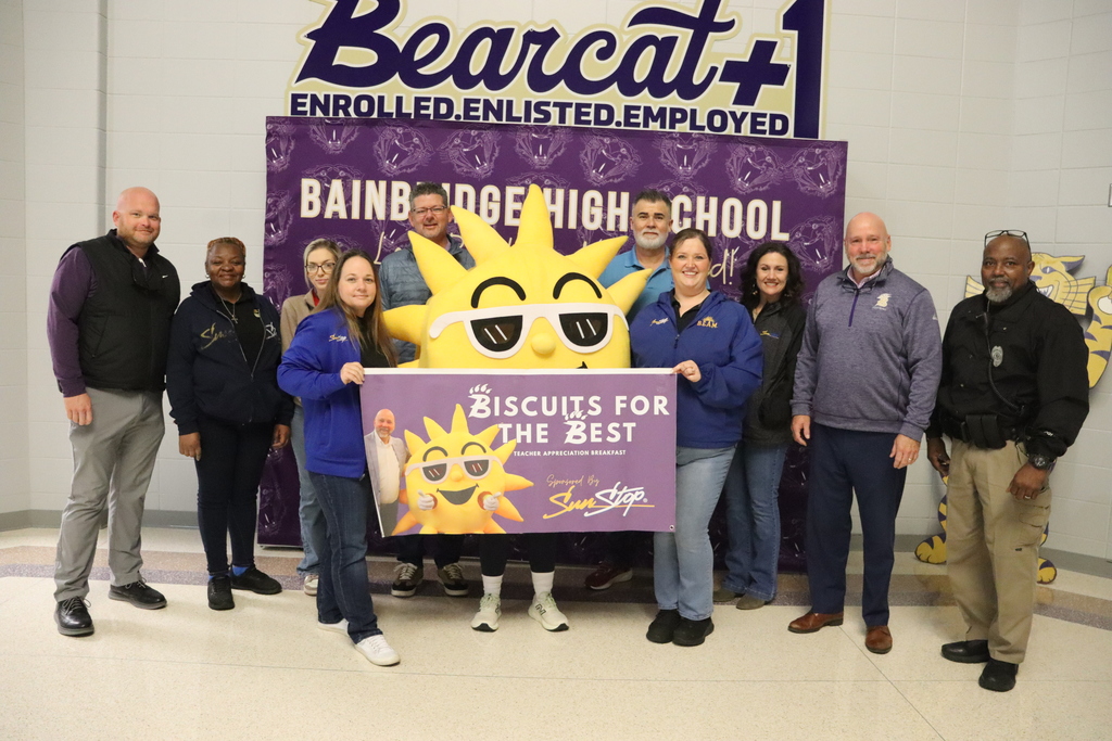 A group of Bainbridge High School staff and community partners pose in front of a school backdrop with the SunStop mascot, holding a “Biscuits for the Best” teacher appreciation banner.