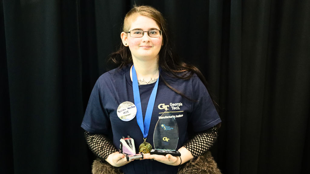 Student wearing a Georgia Tech Manufacturing Institute shirt holds a medal and award plaque in front of a black backdrop.