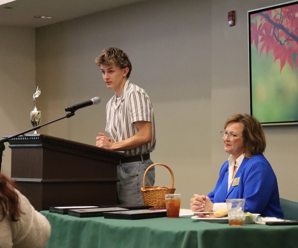 Mitchell Carter speaking at a podium during a recognition event, with two adults seated beside him at a table.