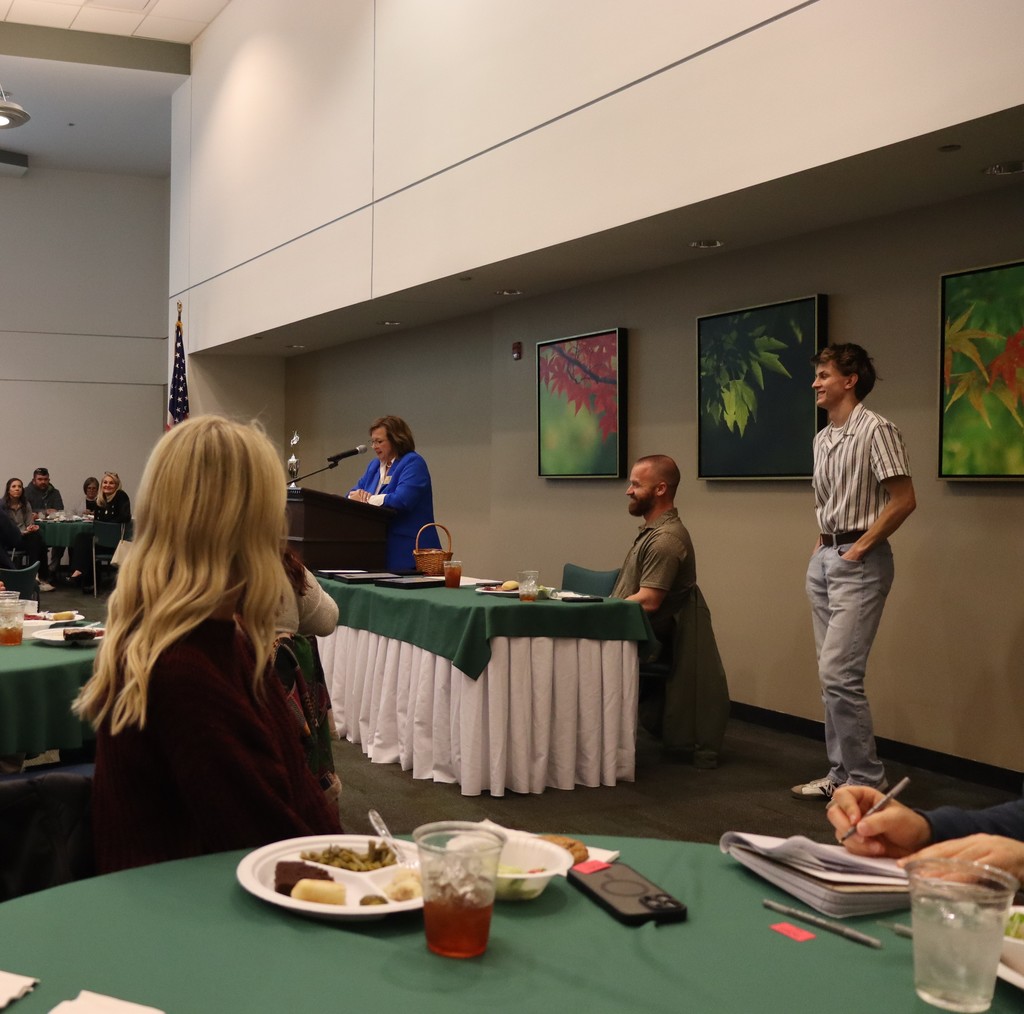 Mitchell Carter speaking at a podium during a recognition event, with two adults seated beside him at a table.