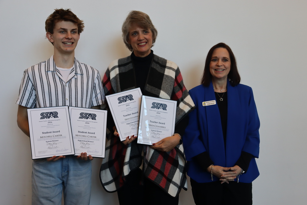 Mitchell Carter holding two STAR Student Award certificates beside Dr. Heidi Chambers holding two STAR Teacher Award certificates, standing with another woman against a white wall.