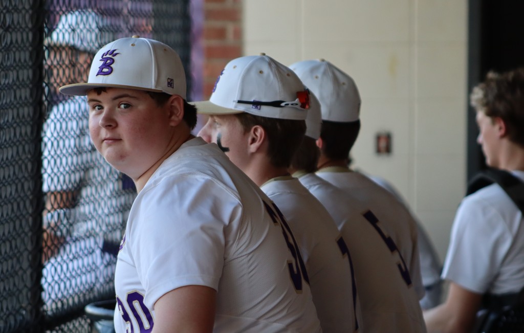   Baseball players in white uniforms lean on the dugout fence, watching the game.