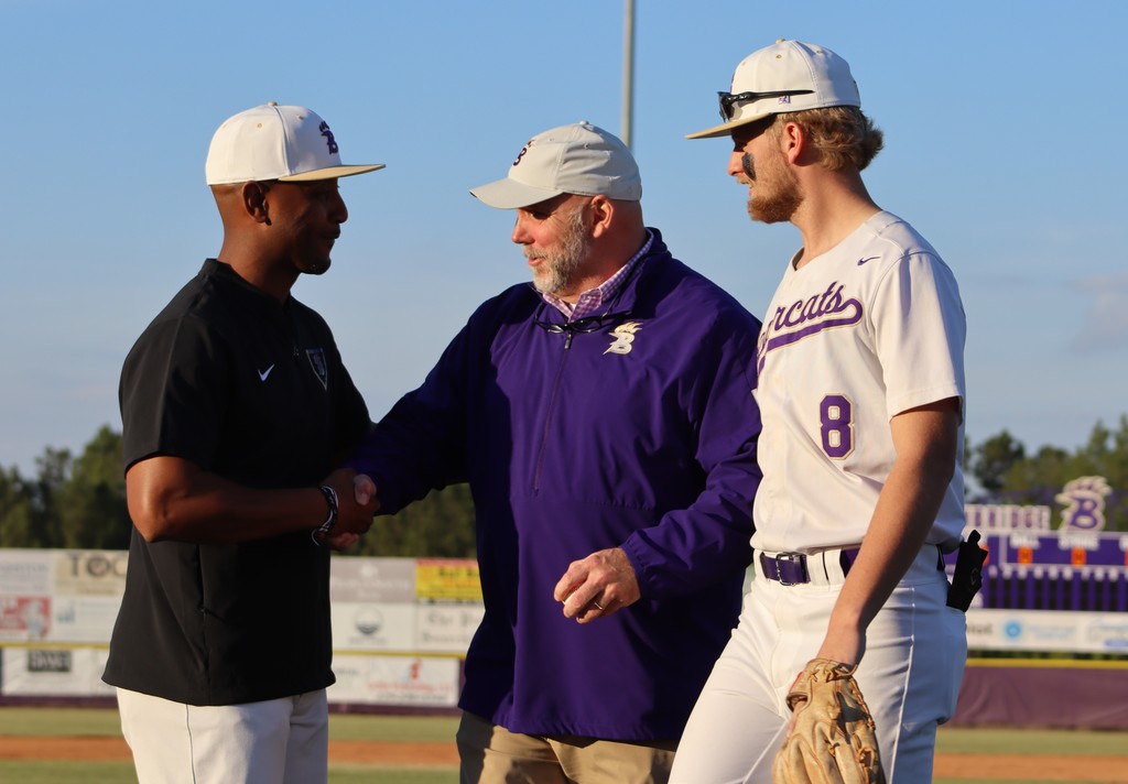 Two coaches shake hands on the field while a player looks on.