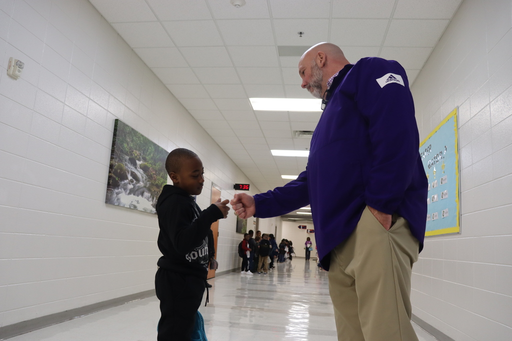 A school administrator in a purple pullover gives a fist bump to a young student in a hallway as other students line up in the background.