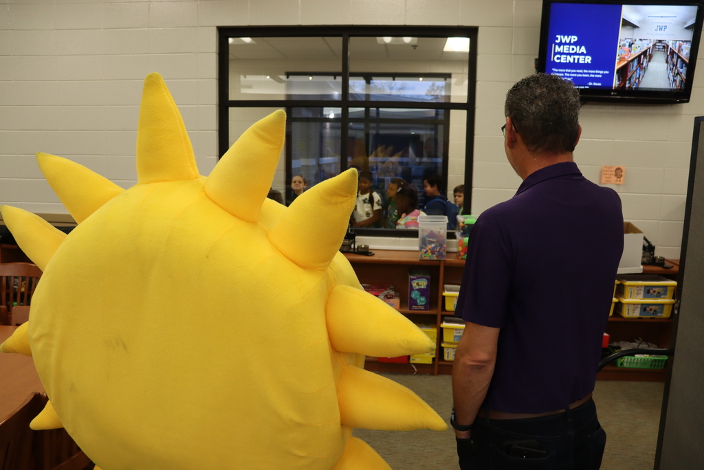A large yellow sun mascot and a staff member stand facing a window inside the media center, looking toward a group of students gathered outside.