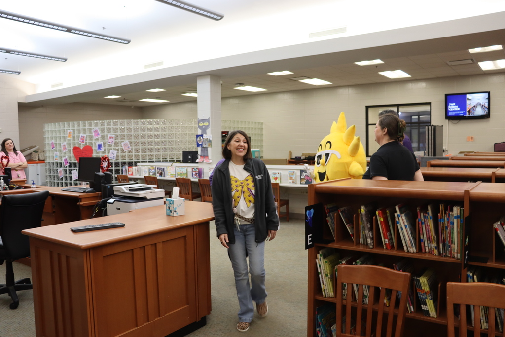 A smiling staff member walks through the media center as others talk in the background near bookshelves and a large sun mascot character.